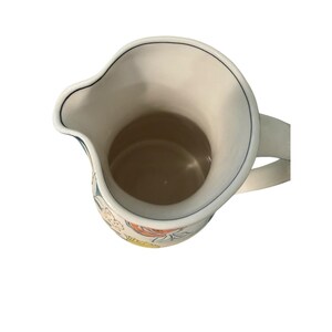 May include: A white ceramic pitcher with a curved spout and a handle. The pitcher has a floral design in shades of orange, yellow, and blue. A thin blue line trims the rim.