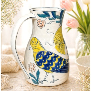 May include: White ceramic pitcher with a large handle, decorated with a yellow and blue bird design, pink flowers, and green leaves. The pitcher has a wide opening and a curved spout, perfect for serving beverages.