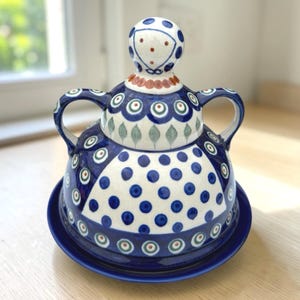 Pode incluir: Uma manteigueira de cerâmica azul e branca com padrão de bolinhas. A tampa tem uma pega decorativa e duas pegas laterais. A base é um prato azul a condizer. O prato é decorado com um estilo de arte popular. Cerca de 15 cm de diâmetro.