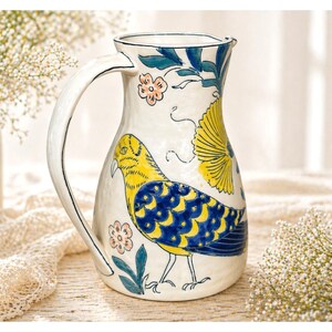 May include: A white ceramic pitcher with a large handle, decorated with a yellow and blue bird design. The pitcher also features floral and leaf accents in blue and pink. The pitcher is a decorative item.