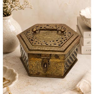 May include: An antique brass hexagonal box with intricate floral and geometric cut-out designs. The box features a hinged lid with a handle and a clasp closure. The box is likely used for storing jewelry or small keepsakes.