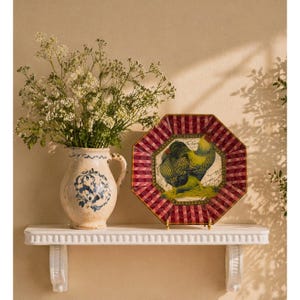 May include: A decorative plate with a rooster design, set on a white shelf. The plate has a red and white striped border. Beside it is a cream-colored pitcher with blue floral patterns, holding white flowers. The shelf is supported by white brackets.