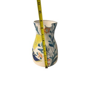 May include: Cream-colored ceramic pitcher with a floral design. The pitcher features yellow, blue, and orange flowers and leaves. The pitcher is approximately 10 inches tall.