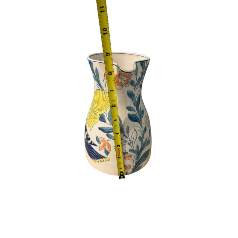 May include: Cream-colored ceramic pitcher with a floral design. The pitcher features yellow, blue, and orange flowers and leaves. The pitcher is approximately 10 inches tall.