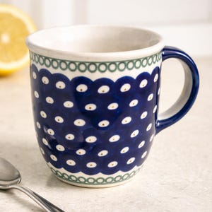 May include: A ceramic mug with a dark blue background and a white polka dot pattern. The rim is decorated with a green and white scalloped design. The handle is dark blue, and the interior is white.