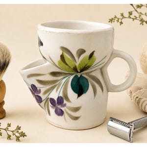 May include: A white ceramic shaving scuttle with a side spout and a handle, featuring hand-painted floral motifs in shades of green, teal, and purple. The vessel has a wide top opening and a cylindrical base, designed for holding shaving lather.