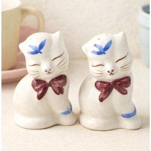 Può includere: Un paio di saliere e pepiere a forma di gatto in ceramica bianca. Ogni gatto ha un papillon bordeaux, dettagli blu e occhi chiusi. I condimenti sono decorati con disegni floreali blu sulla testa.