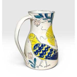 May include: White ceramic pitcher with a curved handle, decorated with a yellow and blue bird design, pink flowers, and blue leaves. The pitcher has a silver rim and a spout for pouring.