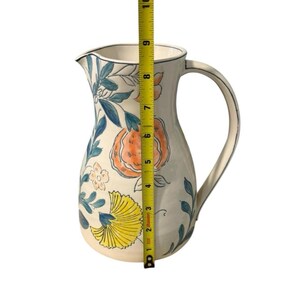 May include: A white ceramic pitcher with a floral design featuring orange, yellow, and blue accents. The pitcher has a curved handle and a spout. The height of the pitcher is approximately 10 inches.