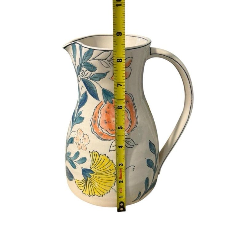 May include: A white ceramic pitcher with a floral design featuring orange, yellow, and blue accents. The pitcher has a curved handle and a spout. The height of the pitcher is approximately 10 inches.