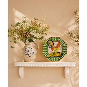 May include: A decorative shelf displays a floral arrangement in a ceramic vase and a decorative plate. The plate features a rooster design with green, yellow, and red accents. The vase has a blue and white floral pattern.