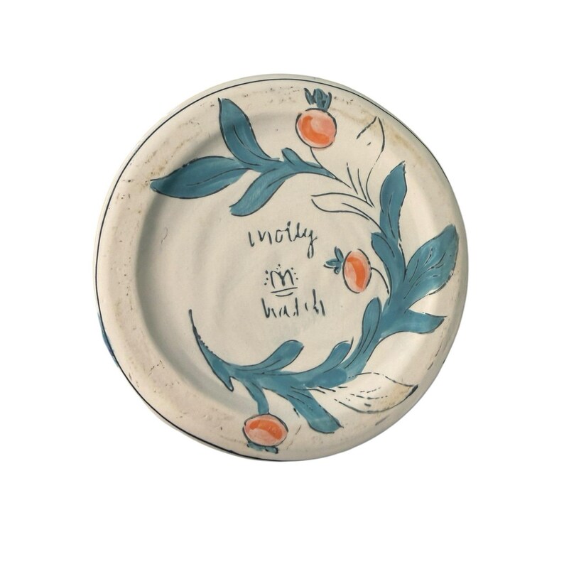May include: A round, off-white ceramic plate with a hand-painted design. The design features teal-colored leaves and orange berries. The plate has text in the center. The rim is outlined in black.