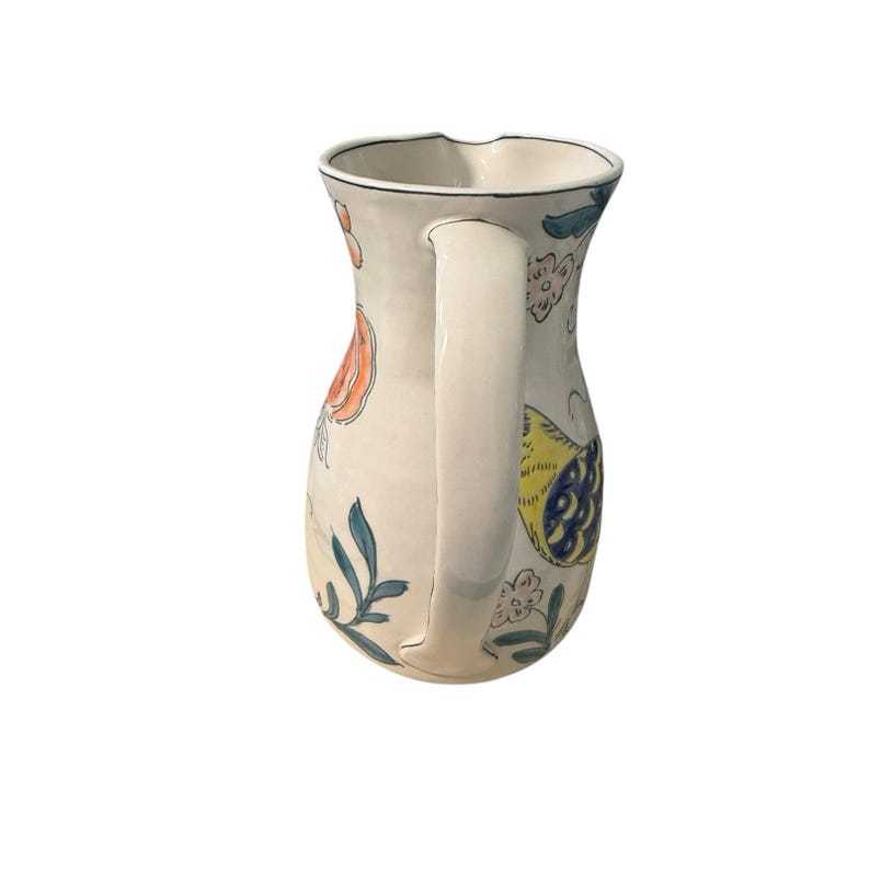 May include: Cream-colored ceramic pitcher with a curved handle and a spout. The pitcher is decorated with colorful floral and leaf designs in orange, blue, and yellow. The rim is outlined with a dark blue line.