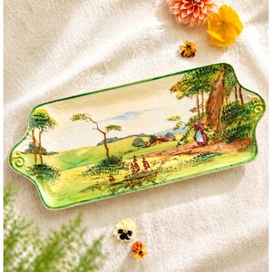 Puede incluir: Bandeja rectangular de cerámica con un paisaje pintado a mano. La bandeja tiene un borde y asas verdes, con una escena campestre en tonos amarillos, verdes y azules. Flores decorativas están dispersas alrededor de la bandeja.