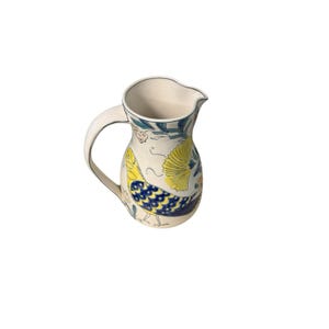 May include: A cream-colored ceramic pitcher with a curved handle and a pouring spout. The pitcher is decorated with a yellow and blue bird design, along with floral and leaf patterns in blue, green, and orange. The rim is outlined in blue.