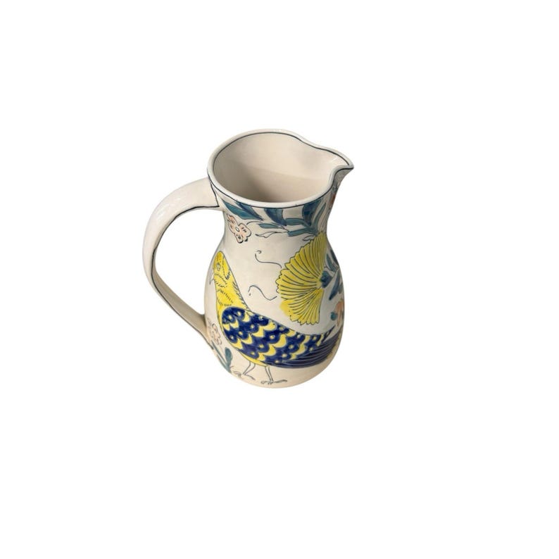May include: A cream-colored ceramic pitcher with a curved handle and a pouring spout. The pitcher is decorated with a yellow and blue bird design, along with floral and leaf patterns in blue, green, and orange. The rim is outlined in blue.