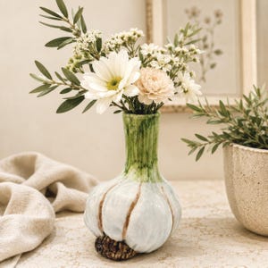 Könnte beinhalten: Eine Keramikvase in Form einer Knoblauchknolle mit weißem, rundem Körper, braunen Wurzeldetails an der Basis und einem grünen Hals. Die Vase enthält einen Strauß aus weißen Gänseblümchen, kleinen weißen Füllblumen und grünen Blattstielen.
