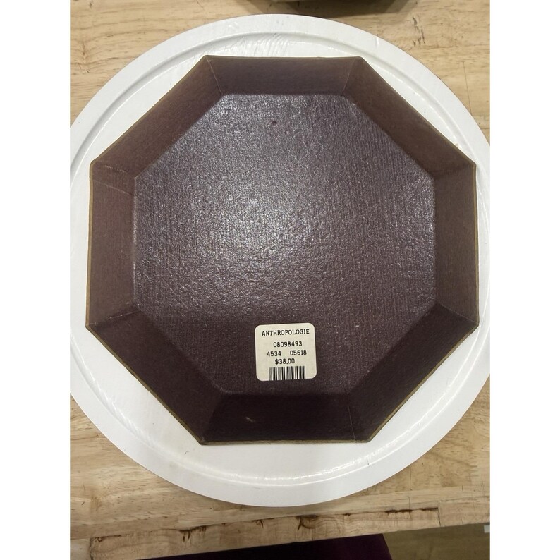 May include: An octagonal brown dish with a textured surface sits inside a white, round plate. The dish has a label with the word "ANTHROPOLOGIE" and other details. The dish is on a wooden surface.