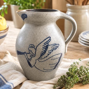 Op de afbeelding: Een gespikkelde lichtgrijze keramische kan met een donkerblauwe, handgeschilderde illustratie van een engel die op een trompet speelt. De kan heeft een rond lichaam, een brede tuit en een gebogen handvat aan de rechterkant met een glazuur.