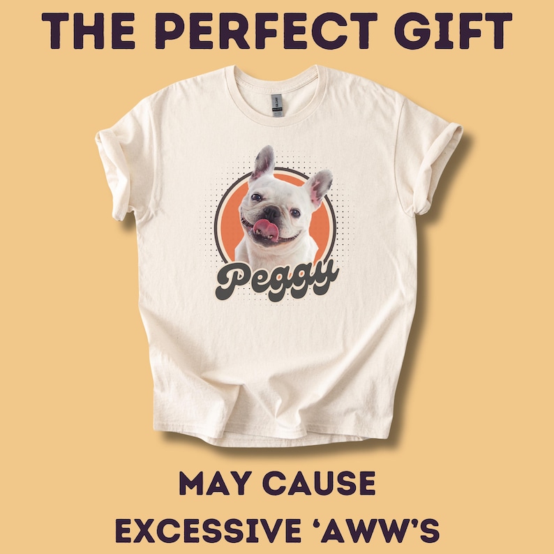 Op de afbeelding: Cr&egrave;mekleurig T-shirt met een lachende witte Franse Bulldog genaamd Peggy. De hond staat in een oranje cirkel. De tekst "THE PERFECT GIFT" en "MAY CAUSE EXCESSIVE 'AWW'S" staan boven en onder de hondengrafiek. Het T-shirt heeft opgerolde mouwen.