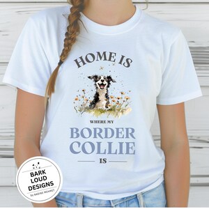 Könnte beinhalten: Weißes T-Shirt mit einer Aquarellillustration eines Border Collies in einem Blumenfeld. Der Text "HOME IS WHERE MY BORDER COLLIE IS" ist oberhalb und unterhalb des Hundes aufgedruckt. Unten befindet sich ein kreisförmiges Logo mit dem Text "BARK LOUD DESIGNS".