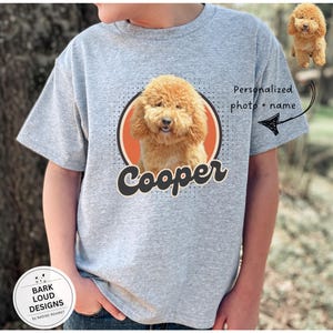 Op de afbeelding: Een grijs t-shirt met een hondenportret in een cirkel, de naam "Cooper" in een scriptlettertype. Het shirt heeft de tekst "Gepersonaliseerde foto + naam" met een pijl die naar de hondenafbeelding wijst. Een kleine hondenafbeelding staat rechtsboven.