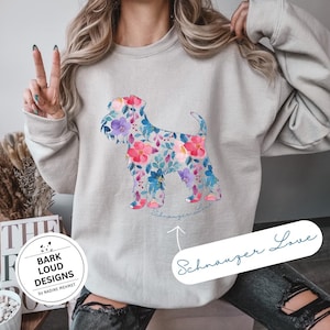 Könnte beinhalten: Beiges Sweatshirt mit floralem Schnauzer-Design. Die Hundesilhouette ist mit rosa, blauen und lila Blumen gefüllt. Die Worte "Schnauzer Love" sind darunter in Kursivschrift geschrieben.