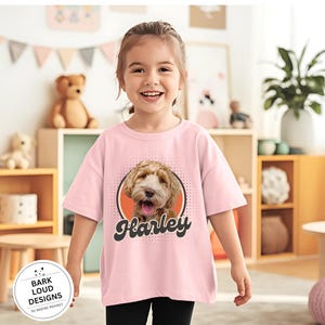 Op de afbeelding: Lichtroze T-shirt met een hondenportret en de naam "Harley" in een retro lettertype. De hond is lichtbruin. Het shirt heeft ook de tekst "Bark Loud Designs by Nadine Memmet" in een cirkelvormig logo.