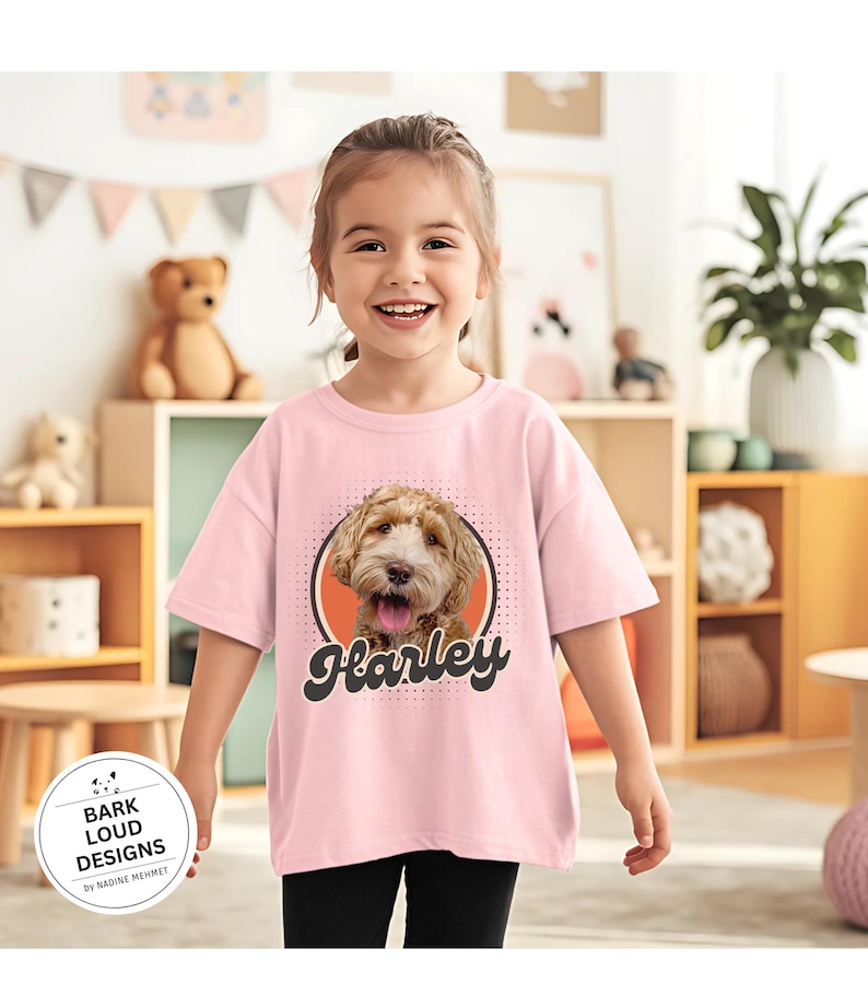 Op de afbeelding: Lichtroze T-shirt met een hondenportret en de naam "Harley" in een retro lettertype. De hond is lichtbruin. Het shirt heeft ook de tekst "Bark Loud Designs by Nadine Memmet" in een cirkelvormig logo.