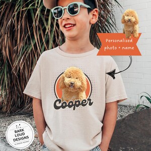 Op de afbeelding: Beige t-shirt met een cartoonafbeelding van een goldendoodle hond en de naam "Cooper" in zwart script. Het ontwerp bevat een oranje cirkel en de tekst "Gepersonaliseerde foto + naam".