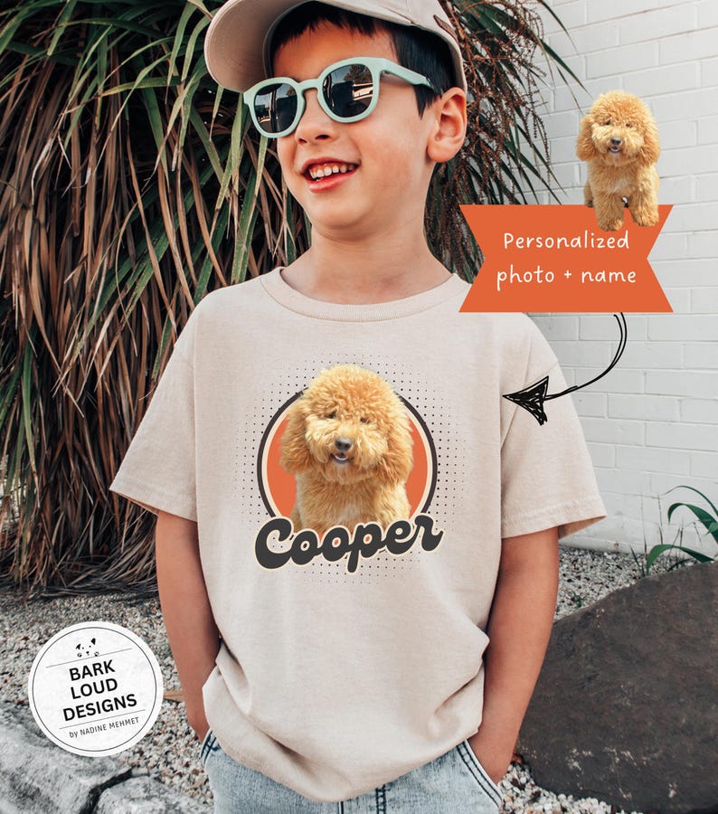 Op de afbeelding: Beige t-shirt met een cartoonafbeelding van een goldendoodle hond en de naam "Cooper" in zwart script. Het ontwerp bevat een oranje cirkel en de tekst "Gepersonaliseerde foto + naam".