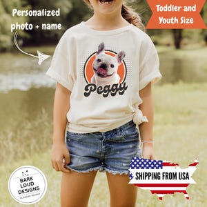 Op de afbeelding: Een cr&egrave;mekleurig T-shirt met een afbeelding van een witte Franse Bulldog met zijn tong uit, in een oranje cirkel met een zwarte stippellijn. De naam van de hond, "Peggy", staat eronder in een retro lettertype. Het shirt is gelabeld met "Peuter- en jeugdmaat".