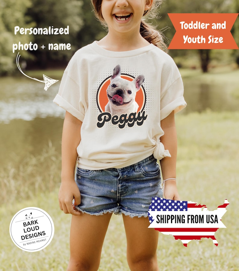 Op de afbeelding: Een cr&egrave;mekleurig T-shirt met een afbeelding van een witte Franse Bulldog met zijn tong uit, in een oranje cirkel met een zwarte stippellijn. De naam van de hond, "Peggy", staat eronder in een retro lettertype. Het shirt is gelabeld met "Peuter- en jeugdmaat".