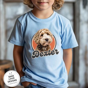 Op de afbeelding: Lichtblauw T-shirt met een hondenportret in een cirkel en de naam "Dexter" in een retro lettertype. De hond heeft bruin en wit haar. Het T-shirt is een T-shirt met korte mouwen en een ronde hals.