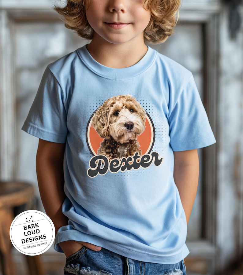 Op de afbeelding: Lichtblauw T-shirt met een hondenportret in een cirkel en de naam "Dexter" in een retro lettertype. De hond heeft bruin en wit haar. Het T-shirt is een T-shirt met korte mouwen en een ronde hals.