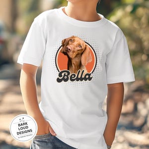 Op de afbeelding: Wit T-shirt met een bruin hondenportret in een oranje cirkel, de naam "Bella" in een retro lettertype. Het shirt heeft ook een klein logo met de tekst "Bark Loud Designs by Nadine Mehmet".