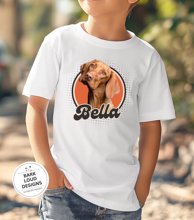 Op de afbeelding: Wit T-shirt met een bruin hondenportret in een oranje cirkel, de naam "Bella" in een retro lettertype. Het shirt heeft ook een klein logo met de tekst "Bark Loud Designs by Nadine Mehmet".