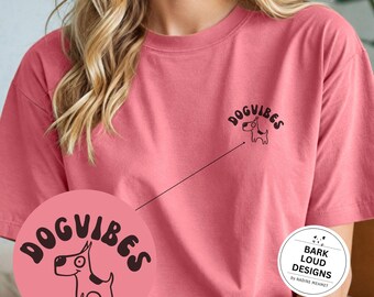 May include: A pink t-shirt with a black graphic of a dog and the text "Dog Vibes". The graphic is on the left side of the shirt. The shirt also has a small white circle on the right side with the text "Bark Loud Designs by Nadine Mennet".