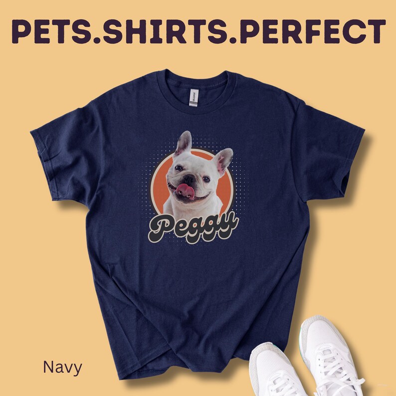 Op de afbeelding: Marineblauw t-shirt met een afbeelding van een witte Franse Bulldog met zijn tong uit, in een oranje cirkel. De naam van de hond, "Peggy", staat eronder. De tekst "PETS.SHIRTS.PERFECT" staat bovenaan.