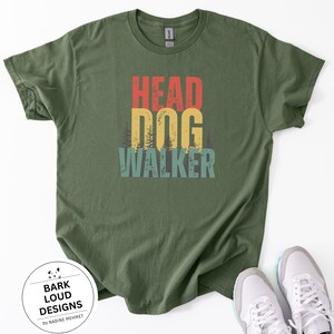 Könnte beinhalten: Olivgrünes T-Shirt mit dem Text "HEAD DOG WALKER" in Rot, Gelb und Türkis. Das Shirt hat einen Vintage-Look. Weiße Turnschuhe sind in der unteren rechten Ecke zu sehen. Ein rundes Logo mit dem Text "BARK LOUD DESIGNS by NADINE MEHMET" befindet sich unten links.