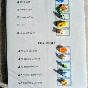 May include: A white paper with the words "SENTENCES" and "ORACIONES" printed on it. The paper has check boxes and miniature toy objects, including a blue hippo, a bag of Cheetos, and a slice of orange. Educational material.