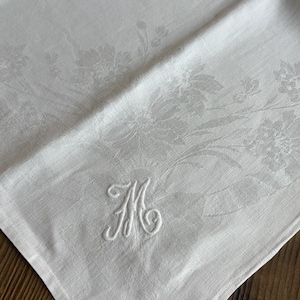 May include: A white linen napkin featuring a tonal damask floral pattern and an embroidered monogram letter M in the corner. The fabric has a subtle sheen and a woven texture, with the stitched letter M standing out in raised white thread.