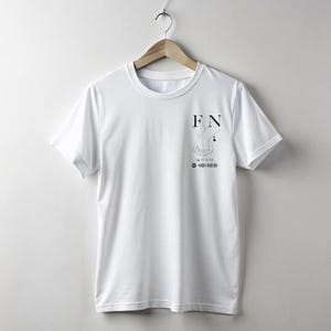 Peut inclure: T-shirt blanc à col rond, suspendu à un cintre en bois. Le t-shirt présente un graphisme noir sur le haut de la poitrine gauche avec les lettres "FN", un dessin au trait de mains, une date et un code-barres.
