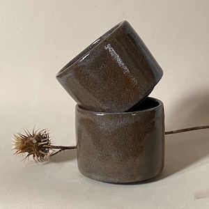 Op de afbeelding: Twee gestapelde, handgemaakte keramische bekers met een donkerbruin, gespikkeld glazuur. De bekers hebben een cilindrische vorm met een licht uitlopende rand. Een gedroogde distel voegt een natuurlijk accent toe.