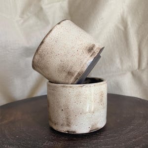 Può includere: Due tazze in ceramica fatte a mano impilate l'una sull'altra. Le tazze sono cilindriche con una glassa bianco sporco macchiettata e accenti marrone scuro sul bordo. L'interno delle tazze è scuro. Le tazze sono su una superficie marrone scuro.
