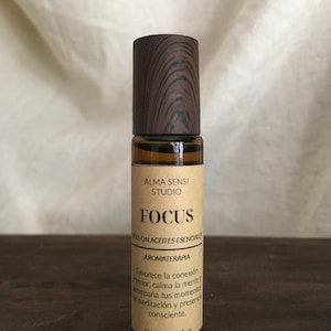 Op de afbeelding: Bruine glazen roll-on fles met een houten dop. Het etiket luidt "FOCUS" en "ROLL-ON ACEITES ESENCIALES". De fles bevat 10 ml aromatherapie-olie. Het etiket bevat ook tekst in het Spaans.