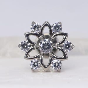 Peut inclure: Boucle d'oreille argentée en forme de fleur, ornée de huit pierres précieuses rondes et transparentes. La boucle d'oreille présente une pierre centrale entourée de sept pierres plus petites, créant un motif floral. Le métal est poli et brillant.