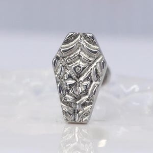 May include: A silver ring with a coffin-shaped face. The ring's face is intricately carved with a symmetrical pattern featuring a central insect-like design. The ring has a polished finish and is set against a soft, neutral background.