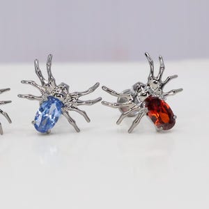 May include: A pair of silver spider earrings with blue and red gemstones. The spiders have eight legs and are detailed with intricate designs.