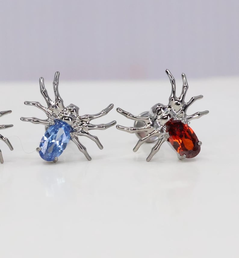 May include: A pair of silver spider earrings with blue and red gemstones. The spiders have eight legs and are detailed with intricate designs.